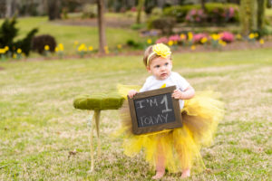 little blonde haired girl standing by green chair at tryphenas garden wedding venue holding sign for one year photography session