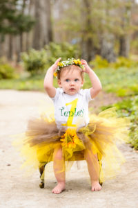 portrait of blonde haired one year old girl sitting in green chair with yellow tutu wearing flowered headband